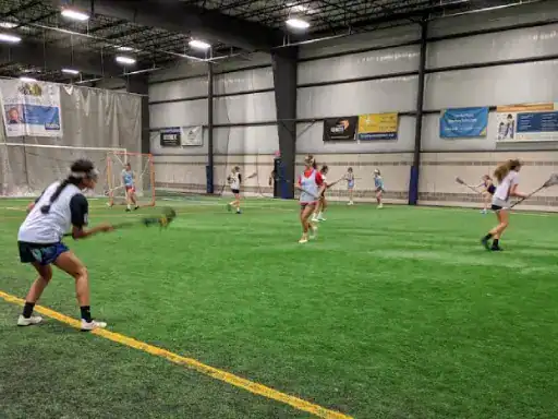 Girls play an indoor lacrosse game on turf under a strong, high ceiling finished by J&P Coatings. The scene shows teamwork and focus as players get ready to score.