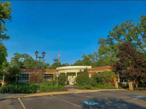 A small library building with big windows sits in a green area. An American flag flies at the entrance. The parking lot has new concrete coatings from J&P Coatings, helping it look clean and cared for.