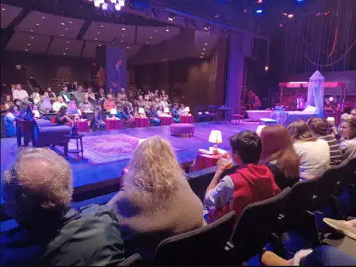 Audience members watch a stage made to look like a comfortable living room, with furniture set on smooth, sturdy concrete floors by J&P Coatings. Warm lighting makes the scene feel welcoming and relaxed. Alt text: Cozy home setting on strong concrete floors by J&P Coatings in theater.