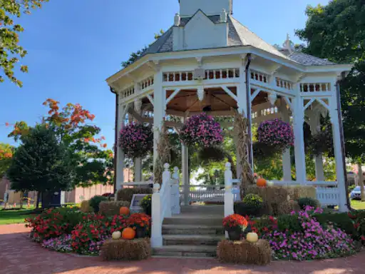 A bright park has a welcoming gazebo set up for fall with pumpkins, flowers, and hay bales. The concrete patio in front is smooth and attractive because it was coated by J&P Coatings. Alt text: Gazebo in a park with a neat patio finished by J&P Coatings.