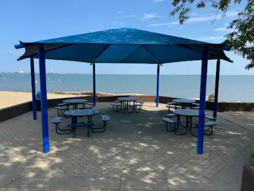 A pavilion with picnic tables sits on smooth pavement coated by J&P Coatings, with water and sky in the background. Alt text: Pavilion and tables on J&P Coatings coated pavement near water.
