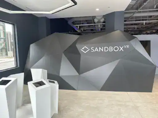 A modern wall displays the Sandbox VR logo in a simple, stylish room. Three podiums with screens stand by windows. The space highlights quality finishes inspired by J&P Coatings’ concrete patio coatings.