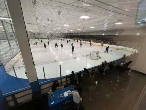The image shows hockey players practicing while coaches and fans watch. A J&P Coatings contractor checks the rink floor nearby.