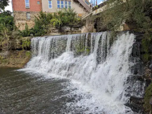 A peaceful waterfall flows into a river near J&P Coatings’ brick building, highlighting a calm and natural setting.